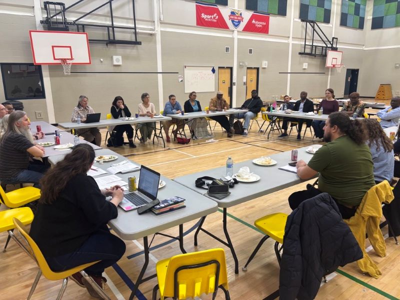 Autour de 4 tables, les représentants et représentantes d'organismes sont assis avec des membres de la communauté de Thompson. La rencontre a lieu dans le gymnase de l'école.